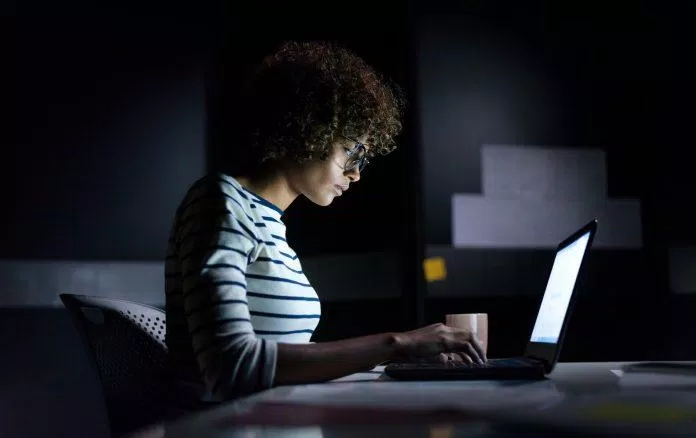 A businesswoman staying late hours in the office concentrating on her work sitting with a laptop and typing.