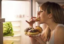Close up image of a young woman with eating disorder, having a midnight snack - eating donuts, in front of the refrigerator.