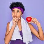 Beautiful african american afro sporty woman doing exercise wearing towel holding heart serious face thinking about question, very confused idea